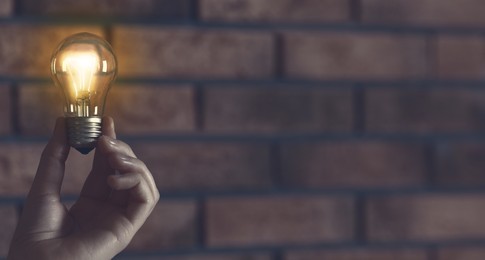 Image of Woman holding glowing light bulb against brick wall, closeup. Idea concept