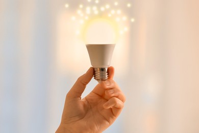 Image of Woman holding glowing light bulb against blurred background, closeup. Idea concept