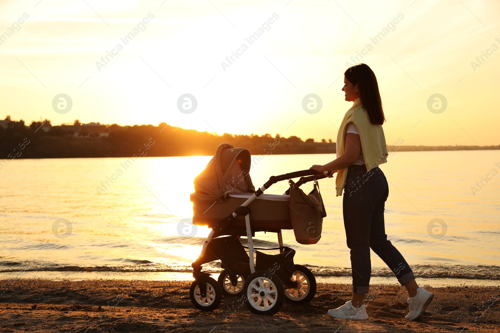 Photo of Happy mother with baby in stroller walking near river at sunset