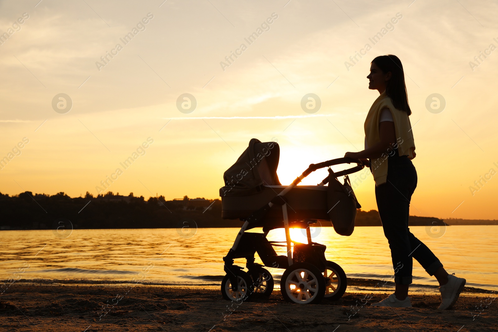 Photo of Happy mother with baby in stroller walking near river at sunset