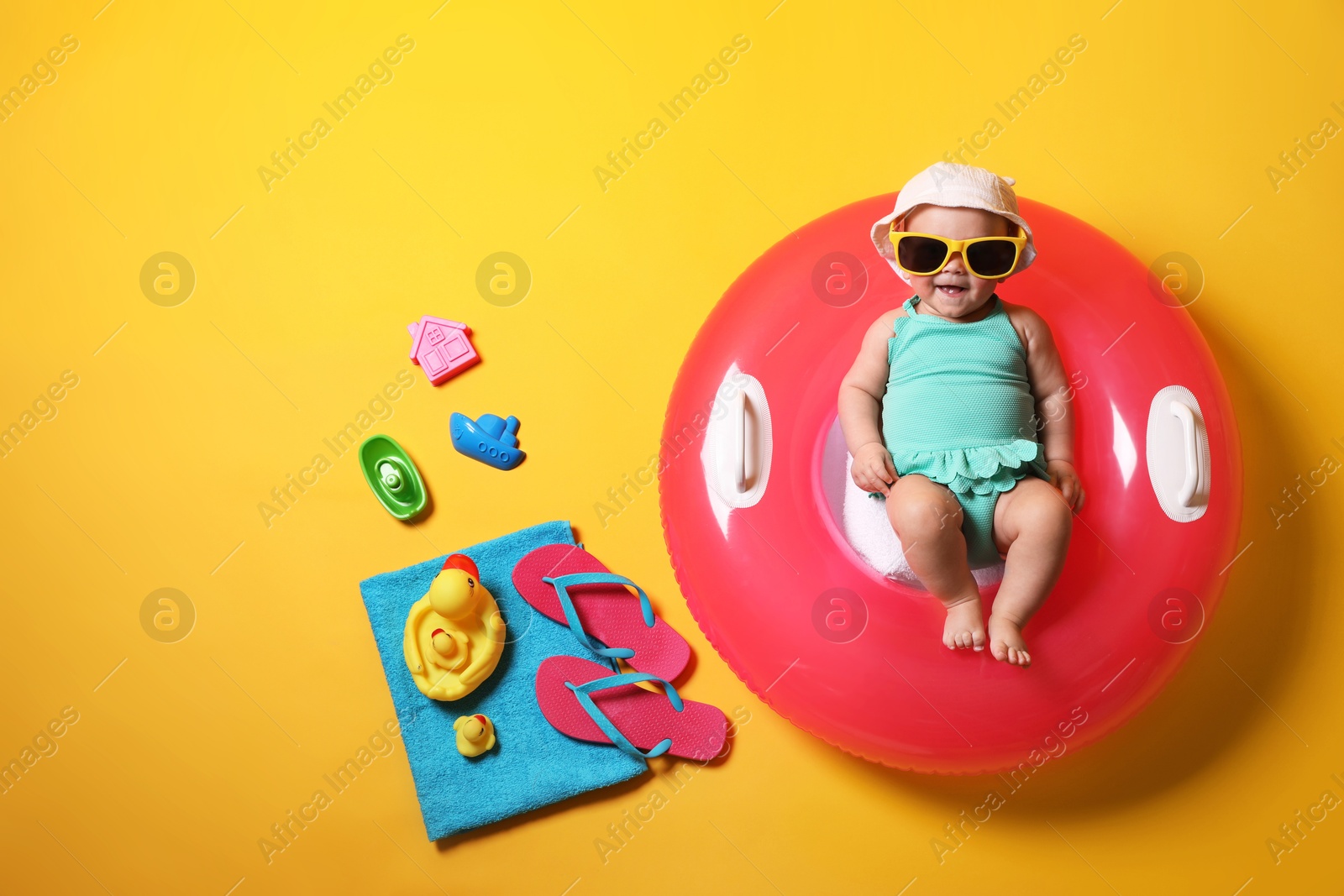 Photo of Cute little baby in sunglasses with inflatable ring and beach accessories on yellow background, top view