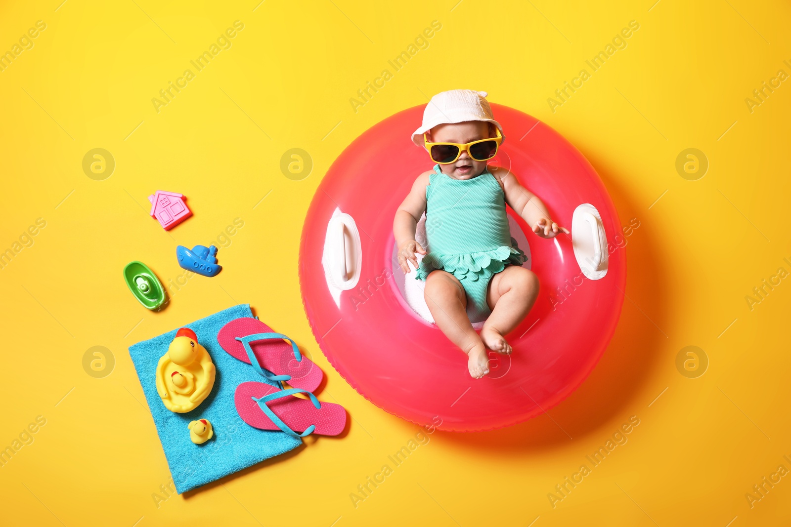 Photo of Cute little baby in sunglasses with inflatable ring and beach accessories on yellow background, top view