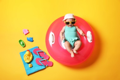Photo of Cute little baby in sunglasses with inflatable ring and beach accessories on yellow background, top view