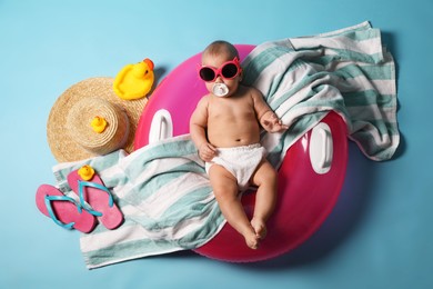 Photo of Cute little baby in sunglasses with inflatable ring and beach accessories on light blue background, top view