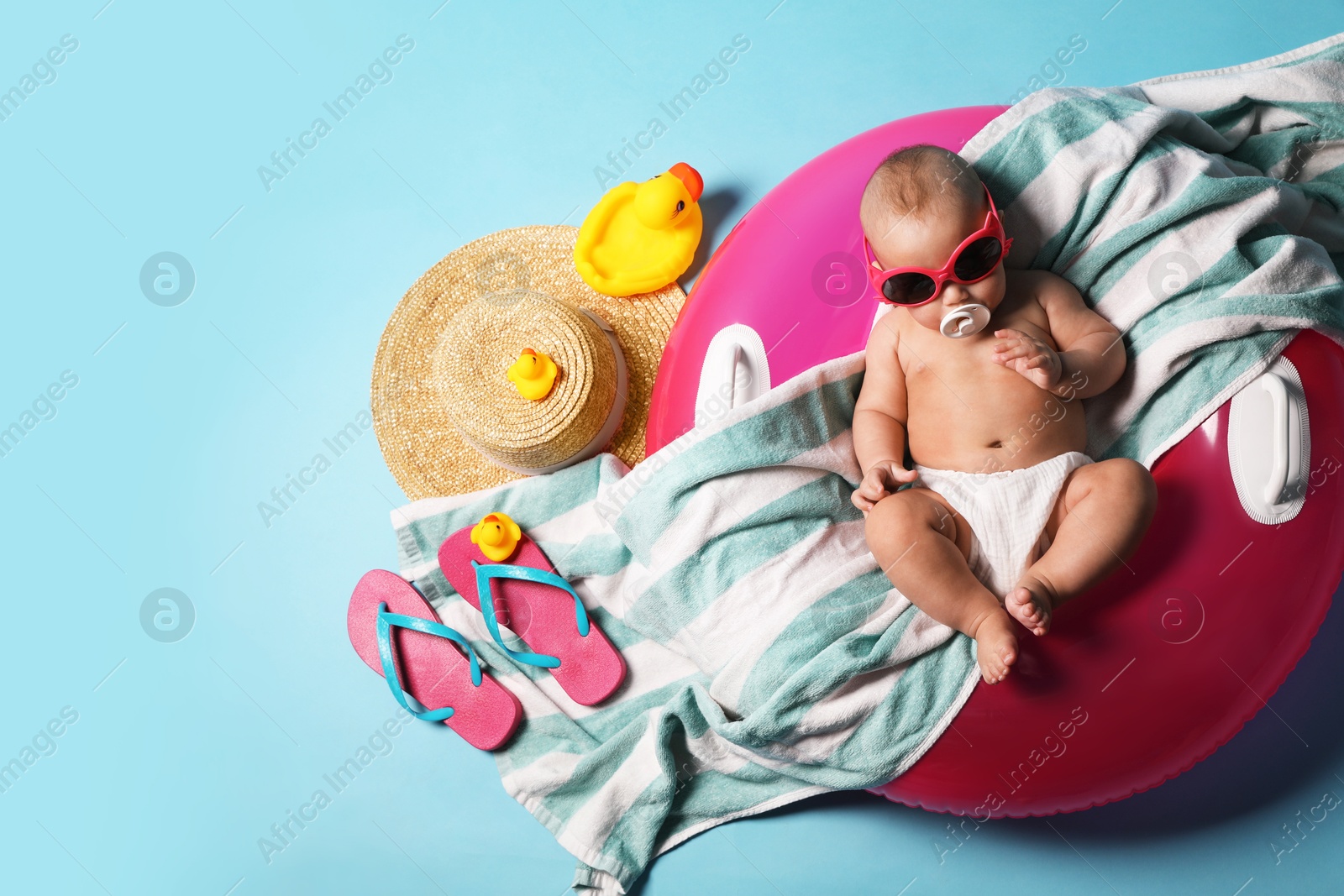 Photo of Cute little baby in sunglasses with inflatable ring and beach accessories on light blue background, top view