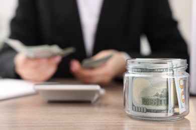 Photo of Woman counting dollars at wooden table indoors, focus on glass jar with money