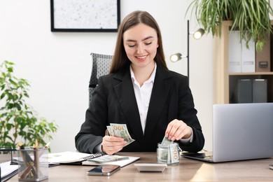 Photo of Woman putting dollars into glass jar at wooden table indoors