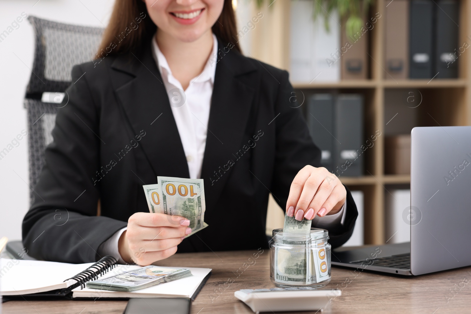 Photo of Woman putting dollars into glass jar at wooden table indoors, closeup