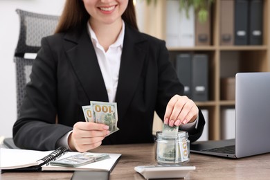 Photo of Woman putting dollars into glass jar at wooden table indoors, closeup