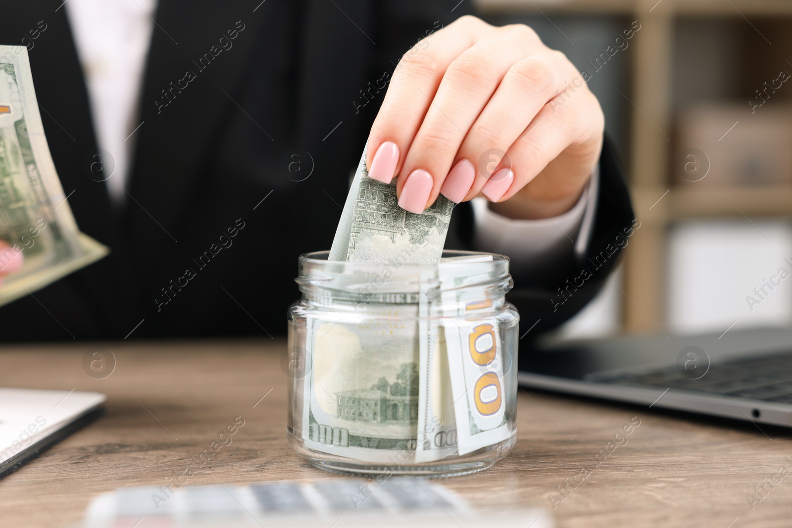 Photo of Woman putting dollars into glass jar at wooden table indoors, closeup
