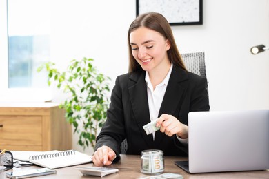 Photo of Woman putting dollars into glass jar and using calculator at wooden table indoors