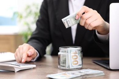 Photo of Woman putting dollars into glass jar and using calculator at wooden table indoors, closeup