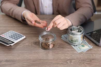 Photo of Woman putting coin into glass jar at wooden table indoors, closeup