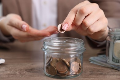 Photo of Woman putting coin into glass jar at wooden table indoors, closeup