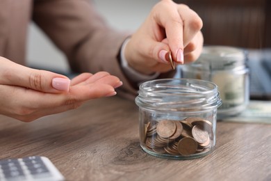 Photo of Woman putting coin into glass jar at wooden table indoors, closeup