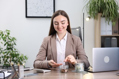 Photo of Woman putting coin into glass jar at wooden table indoors