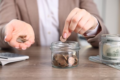 Photo of Woman putting coin into glass jar at wooden table indoors, closeup
