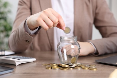 Photo of Woman putting coin into glass jar at wooden table indoors, closeup