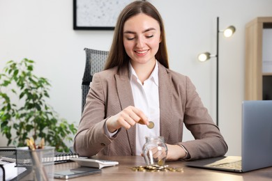 Photo of Woman putting coin into glass jar at wooden table indoors