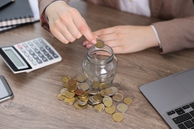 Photo of Woman putting coin into glass jar at wooden table indoors, closeup