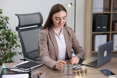 Photo of Woman putting coin into glass jar and using calculator at wooden table indoors