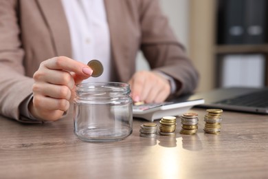 Photo of Woman putting coin into glass jar and using calculator at wooden table indoors, closeup