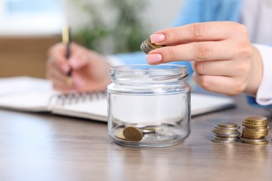 Photo of Woman putting coins into glass jar while working at wooden table indoors, closeup