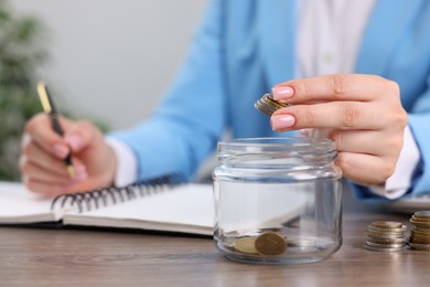 Photo of Woman putting coins into glass jar while working at wooden table indoors, closeup