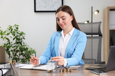 Photo of Woman putting coins into glass jar while working at wooden table indoors