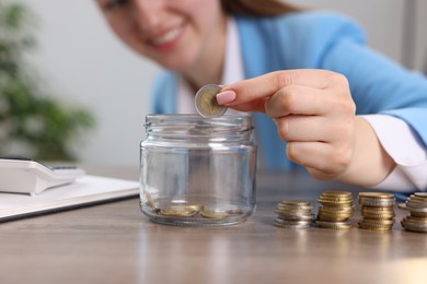 Photo of Woman putting coin into glass jar at wooden table indoors, closeup