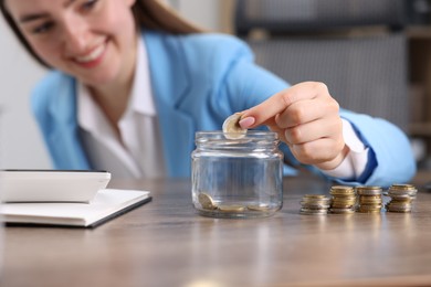 Photo of Woman putting coin into glass jar at wooden table indoors, selective focus