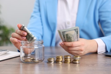 Photo of Woman putting dollar banknote into glass jar at wooden table indoors, closeup
