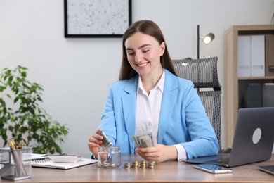 Photo of Woman putting dollar banknote into glass jar at wooden table indoors