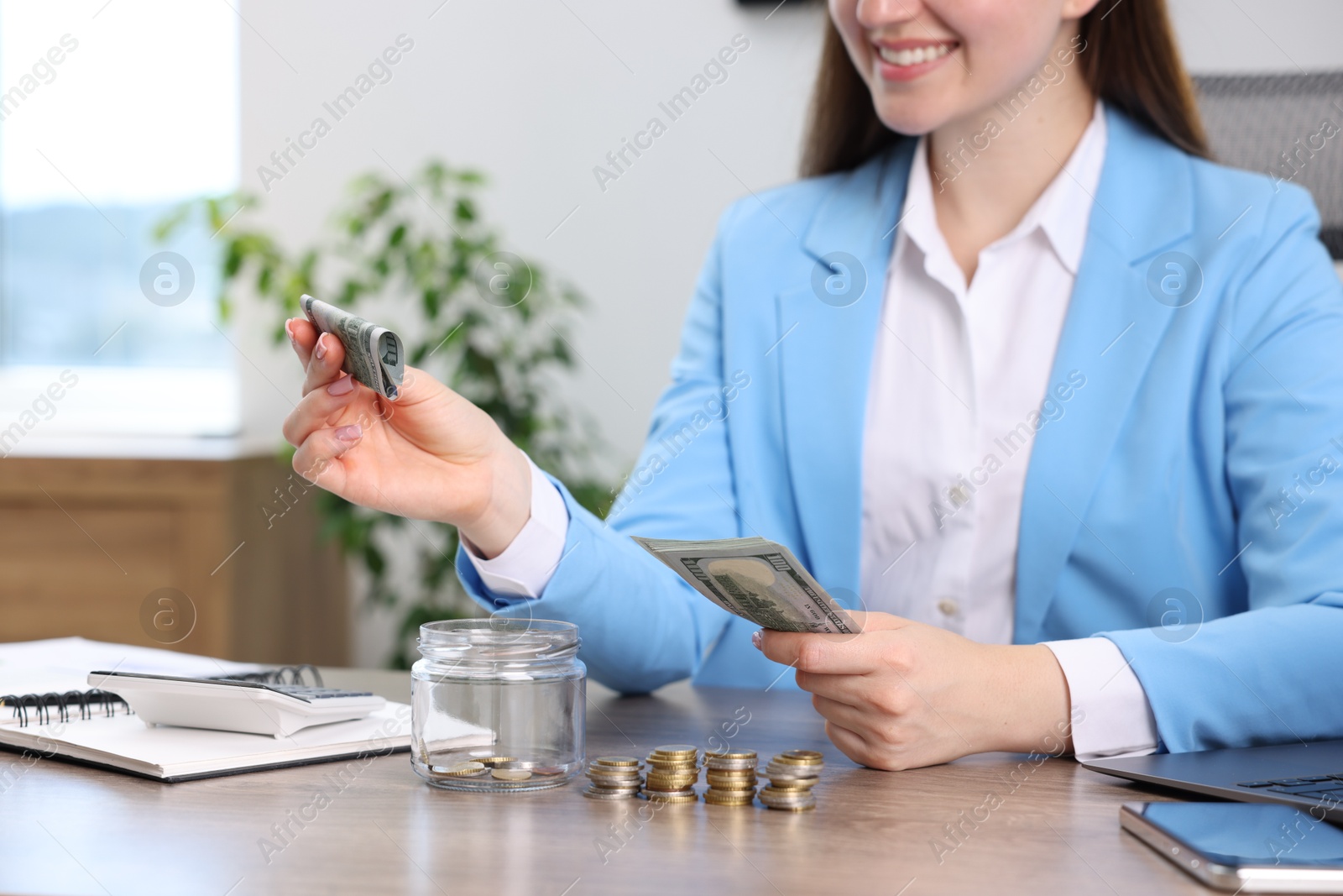 Photo of Woman putting dollar banknote into glass jar at wooden table indoors, closeup
