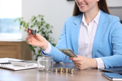 Photo of Woman putting dollar banknote into glass jar at wooden table indoors, closeup