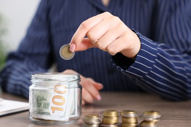 Photo of Woman putting coin into glass jar at wooden table indoors, closeup