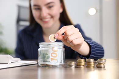 Photo of Woman putting coin into glass jar at wooden table indoors, selective focus
