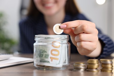 Photo of Woman putting coin into glass jar at wooden table indoors, closeup