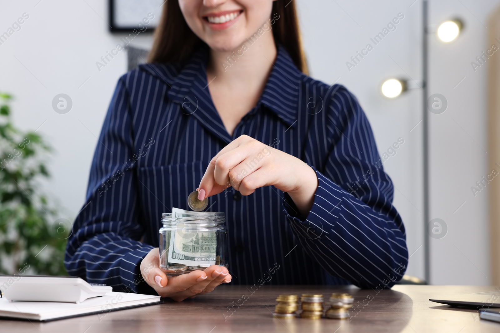 Photo of Woman putting coin into glass jar at wooden table indoors, closeup