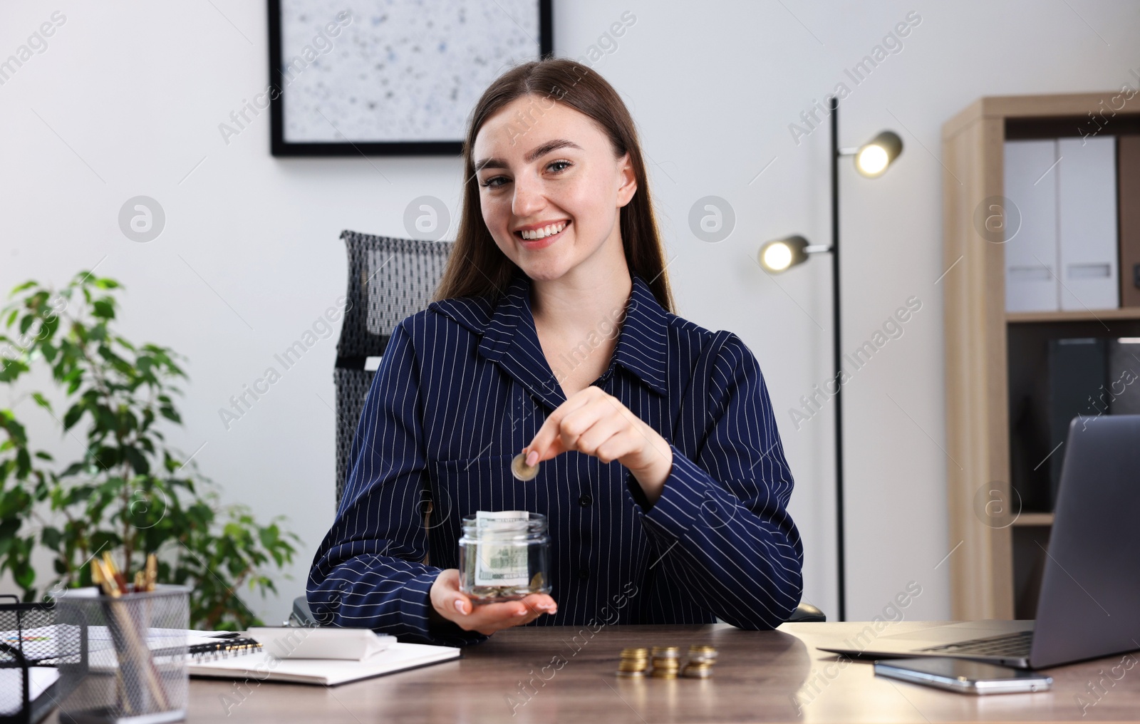Photo of Woman putting coin into glass jar at wooden table indoors