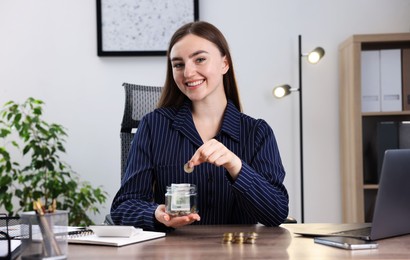 Photo of Woman putting coin into glass jar at wooden table indoors