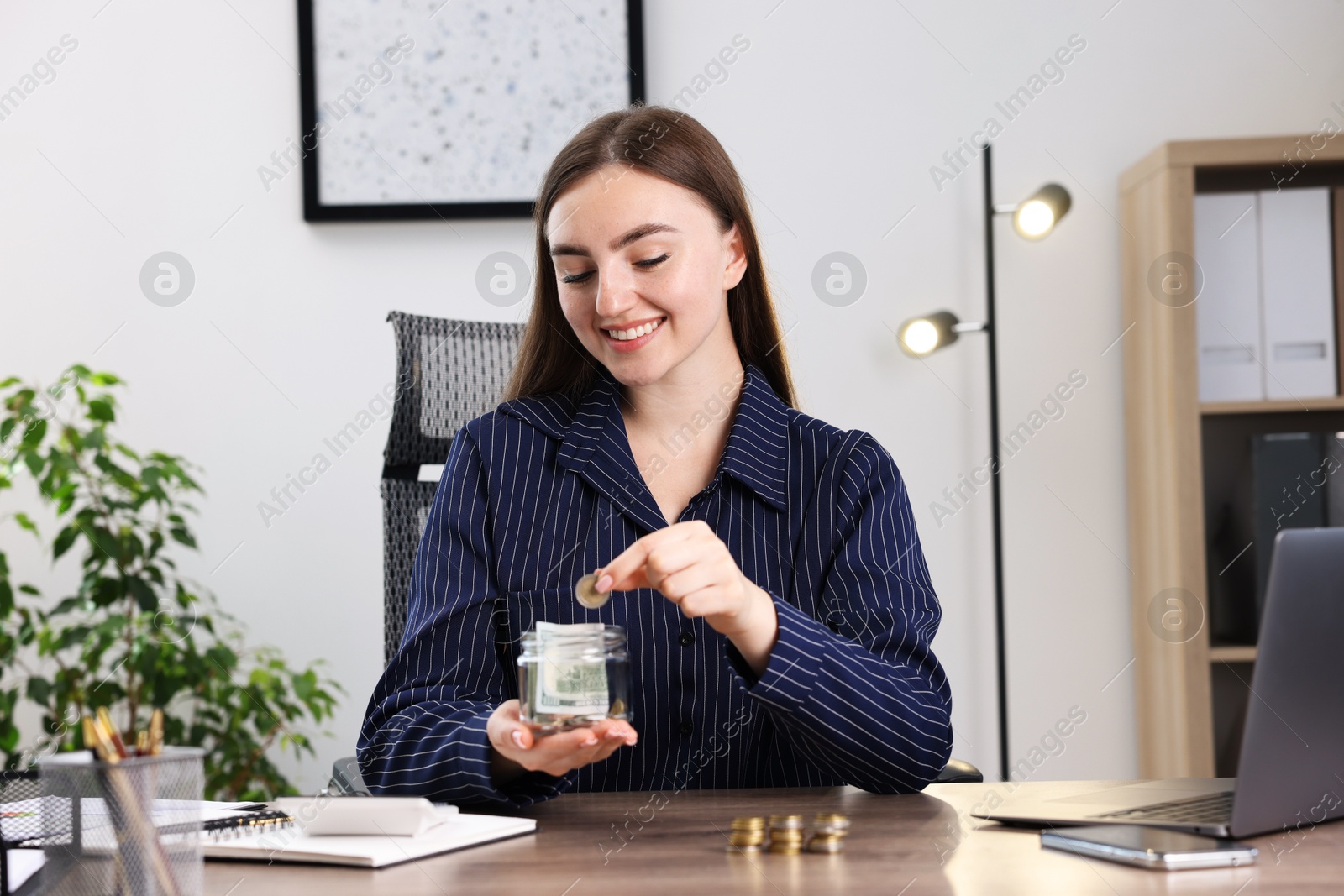 Photo of Woman putting coin into glass jar at wooden table indoors