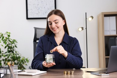 Photo of Woman putting coin into glass jar at wooden table indoors