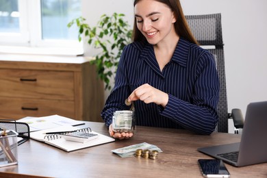 Photo of Woman putting coin into glass jar at wooden table indoors, space for text
