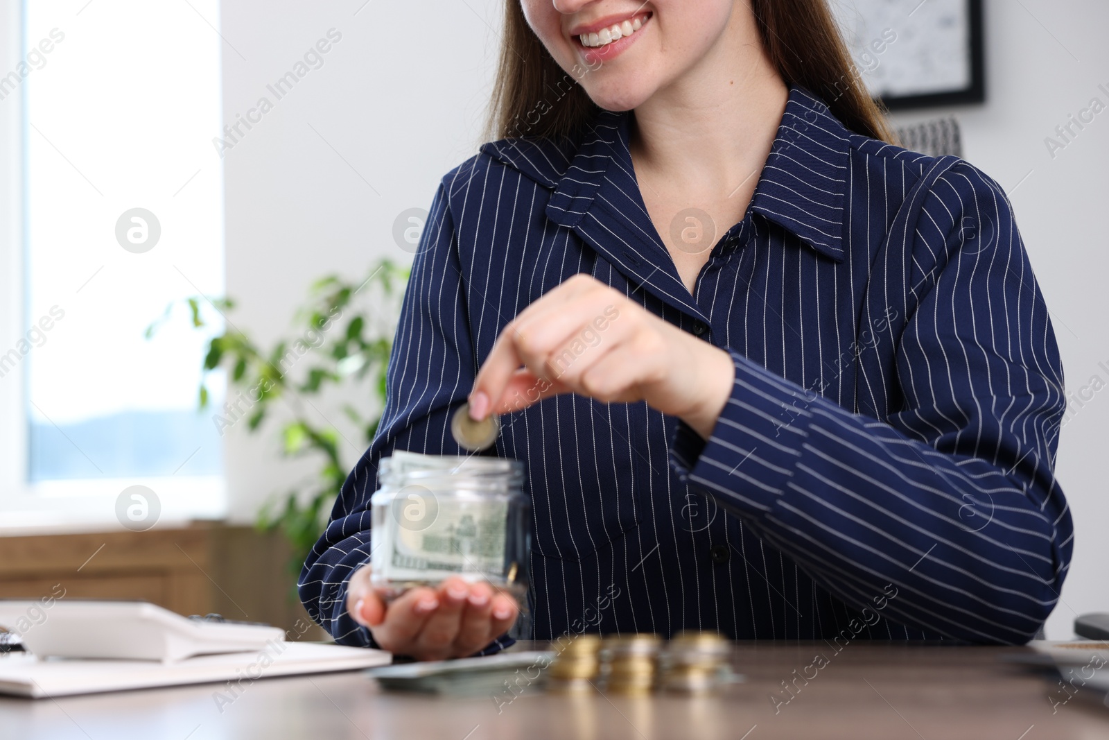 Photo of Woman putting coin into glass jar at wooden table indoors, closeup