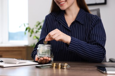 Photo of Woman putting coin into glass jar at wooden table indoors, closeup