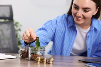 Photo of Woman putting coins into glass jar at wooden table indoors, selective focus