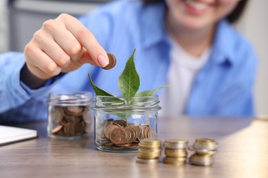 Photo of Woman putting coins into glass jar at wooden table indoors, closeup