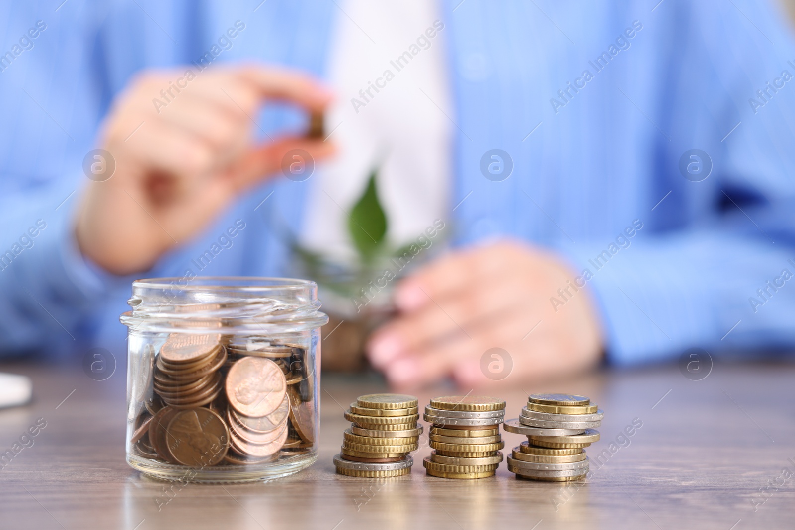 Photo of Woman putting coins into glass jar at wooden table indoors, focus on money