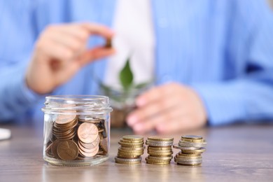 Photo of Woman putting coins into glass jar at wooden table indoors, focus on money